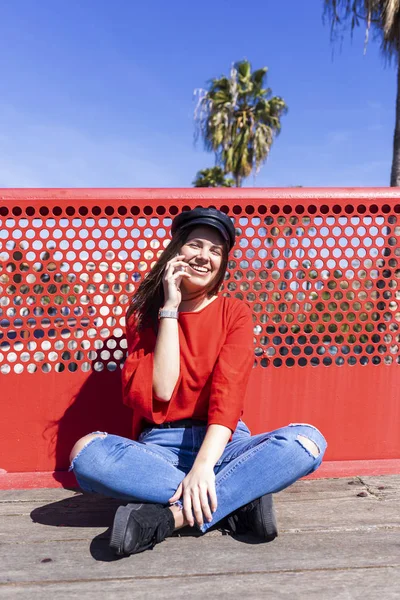 Vista frontal de una hermosa joven vestida con ropa urbana sentada en el suelo mientras usa un teléfono móvil al aire libre en la calle en un día brillante — Foto de Stock