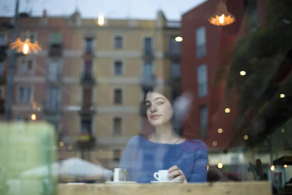 Porträtt av vacker ung dam dricka kaffe och titta på kamera genom glas när du sitter vid bordet i ett kafé — Stockfoto