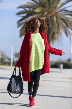 Genç güzel kıvırcık Afro kadının yan görünümü, güneşli bir günde şehir yolundaki ayakta gülümseyen ve uzak bakıyor