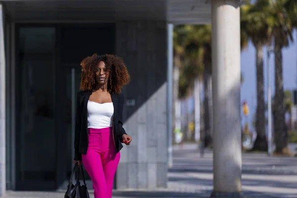 Vista frontale della giovane bella donna riccia che indossa abiti eleganti e borsetta mentre in piedi in strada nella giornata di sole — Foto Stock