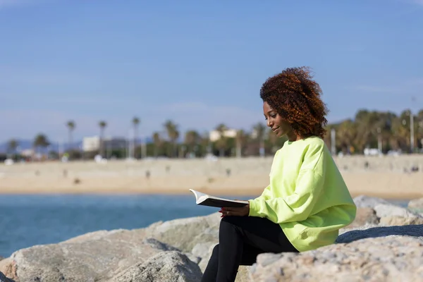 Seitenansicht einer jungen lockigen Afro-Frau, die auf einer Buhne sitzt und ein Buch hält, während sie lächelt und draußen wegschaut — Stockfoto