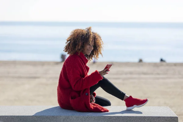 Вид збоку молодий красивий Кучерявий афроамериканець жінка сидить на лавці на пляжі при використанні мобільного телефону на відкритому повітрі — стокове фото
