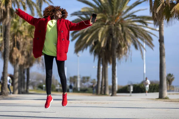 Vista frontal de la joven hermosa rizada mujer afroamericana saltando y bailando mientras escucha música y sonríe al aire libre — Foto de Stock