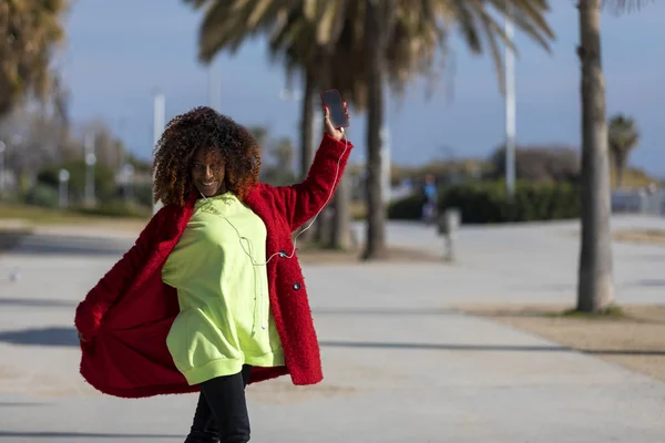 Vista frontal de una joven mujer afro rizada sonriente parada al aire libre mientras sonríe y escucha música con auriculares en un día soleado — Foto de Stock