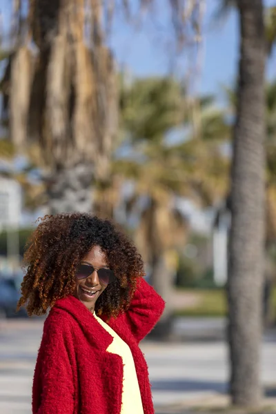 Vista frontal de una joven hermosa mujer afro rizada con gafas de sol y chaqueta roja de pie en una calle de la ciudad mientras toca el cabello y sonríe en un día soleado — Foto de Stock