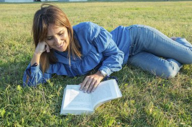 Kitap okurken parkta çim zemin üzerinde yatan gülümseyen bir kadının yan görünümü
