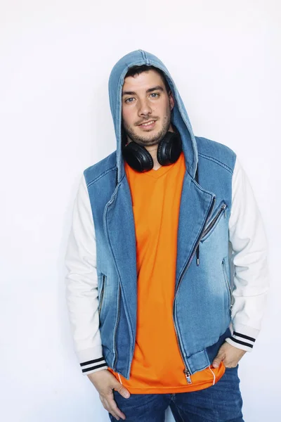 Young handsome man wearing jeans jacket with hood on and headphones on his neck standing against white wall while posing on camera — Stock Photo, Image