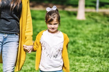 Parkta duran ve güneşli bir günde anne ile el ele tutuşurken kameraya bakan bir baş kravat ile sevimli bir küçük kız ön görünümü