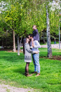 Aile anne, baba ve kızı omuzlarında baba oturur ve veliler gün ışığında parkta öpücük
