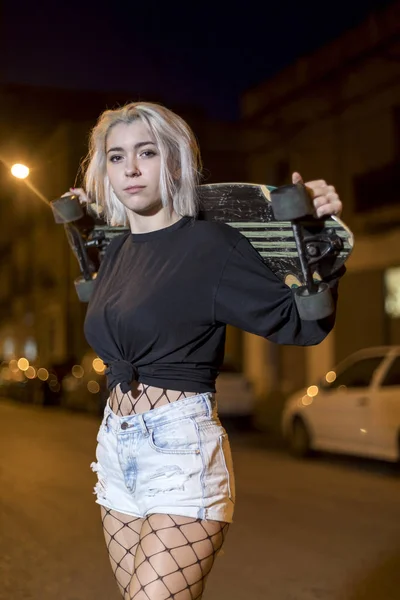 도시에서 밤에 스케이트 보드를 들고 거리에 서있는 동안 반바지에 카메라를 찾고 젊은 예쁜 여자의 전면보기 — 스톡 사진