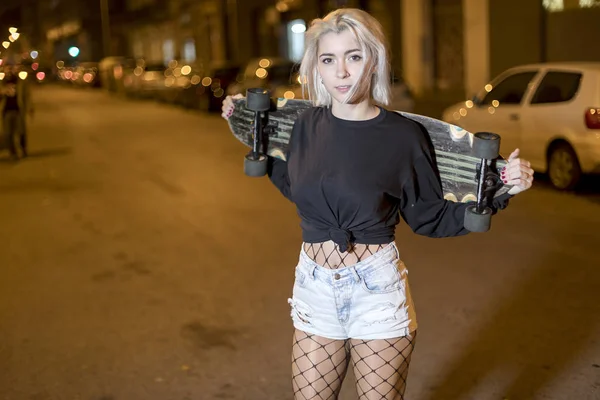 도시에서 밤에 스케이트 보드를 들고 거리에 서있는 동안 반바지에 카메라를 찾고 젊은 예쁜 여자의 전면보기 — 스톡 사진