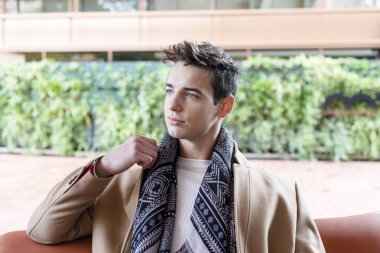 Kışlık kıyafetler giyen genç adam sokakta deri bir bankta oturuyor..