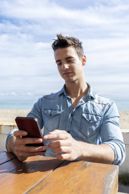 Güneşli bir günde cep telefonu kullanırken bir plaj kulübünde oturan genç adamın ön görünümü