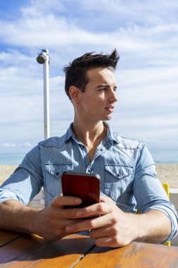 Güneşli bir günde cep telefonu kullanırken bir plaj kulübünde oturan genç adamın ön görünümü