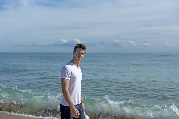 Fiatal vonzó férfi sétál a tengerparton kezében egy pár tornacipő egy napsütéses napon — Stock Fotó