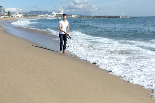 Young attractive man walking on seashore holding a pair of sneakers in a sunny day — Stock Photo, Image