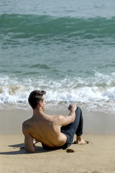 Rückansicht eines jungen, entblößten Mannes in Jeans, der an einem sonnigen Tag am Strand sitzt und wegschaut — Stockfoto