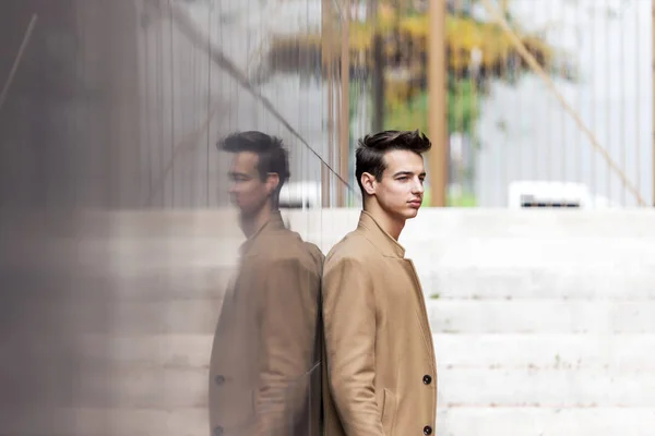 Young handsome man with coat leaning on a reflective wall while looking away on the street — ストック写真