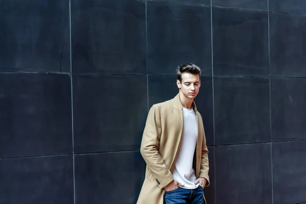 Young man wearing winter clothes in the street while leaning on a black wall and looking down — ストック写真