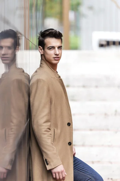 Young handsome man with coat leaning on a reflective wall while looking away on the street — ストック写真