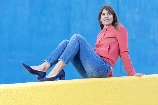 Piękna Dorosła Kobieta w dżinsach leżąca na żółtym ogrodzeniu, patrząc z dala od niebieskiej ściany — Zdjęcie stockowe