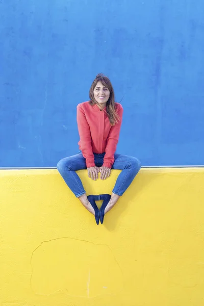 Hermosa mujer adulta en jeans sentados en una valla amarilla mientras mira la cámara contra la pared azul — Foto de Stock