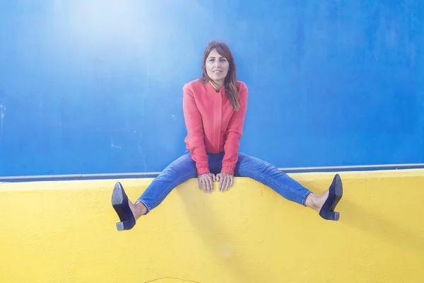 Krásná dospělá žena v džínách, která sedí na žlutém plotu a dívá se na kameru proti modré stěně — Stock fotografie