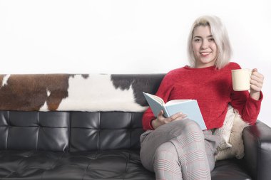Bir fincan çay tutarken kanepeüzerinde kitap okuyan genç mutlu kadın