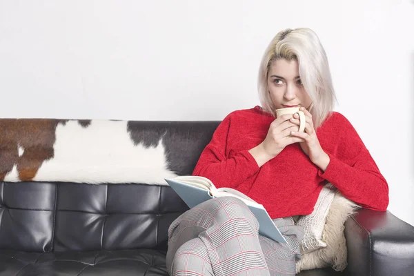 Kavkazská blondýnka sedí na pohovce pije kávu a čte knihu — Stock fotografie