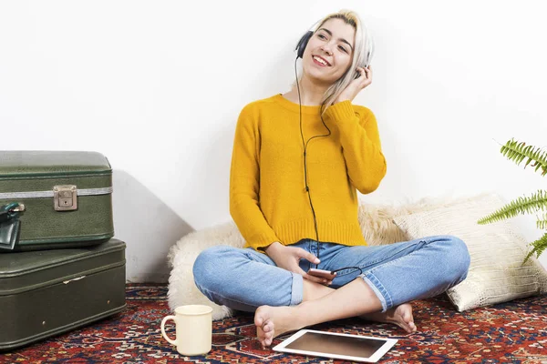 Mujer joven sonriente sentada en la cama con teléfono celular escuchando música con auriculares —  Fotos de Stock
