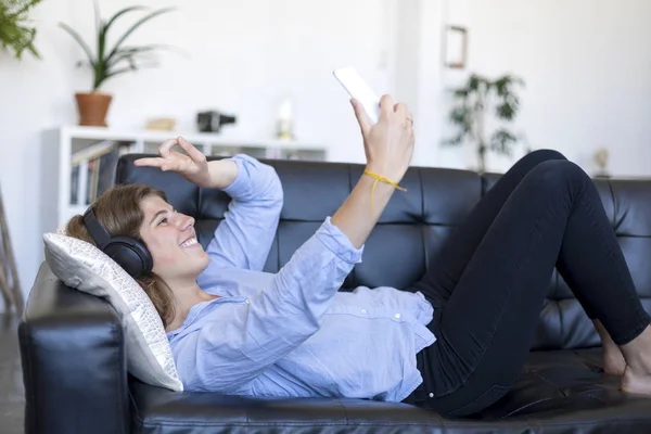 Joven mujer de pelo bastante largo acostada en su espalda en el sofá haciendo selfie en su teléfono — Foto de Stock