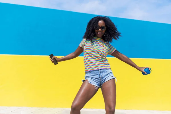 Razza mista sorridente donna nera ritratto con grandi capelli ricci afro contro la parete blu e gialla che balla tenendo in mano uno smartphone — Foto Stock