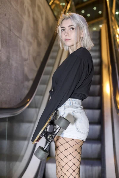 Visão traseira de uma jovem mulher olhando câmera em shorts segurando uma placa longa enquanto está de pé em escadas mecânicas à noite — Fotografia de Stock