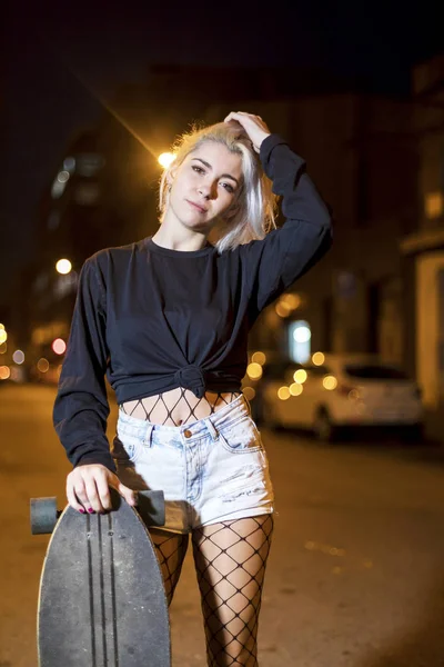 Vue de face de la jeune jolie femme regardant caméra en short tandis que debout dans la rue tenant une planche à roulettes la nuit dans la ville — Photo