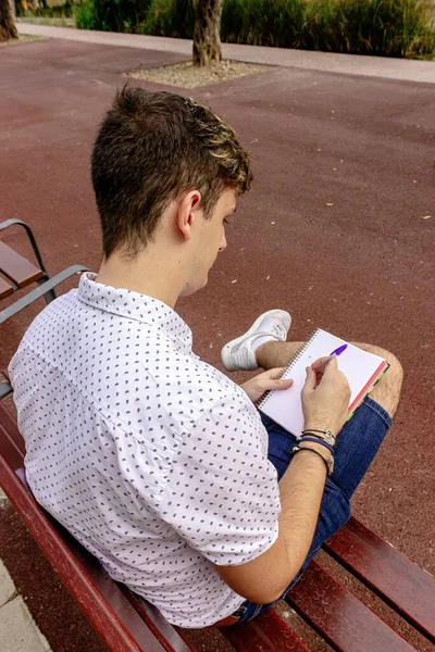 Männlicher Teenager Sitzt Auf Einer Bank Während Einem Notizbuch Schreibt — Stockfoto