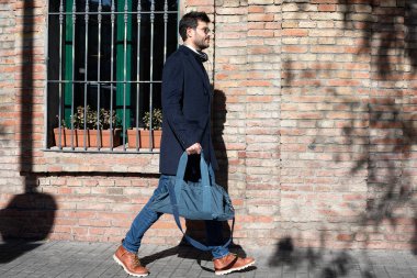 Sakallı, güneş gözlüklü ve günlük giysili bir erkek sokakta yürürken spor çantası taşıyor.