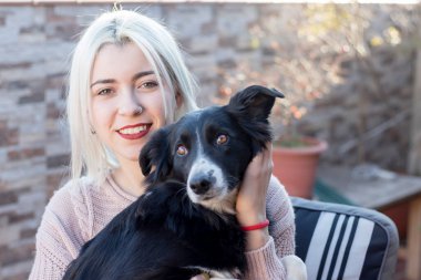 Sarışın genç bir kadın dışarıda terasta otururken köpeğine sarılıyor.