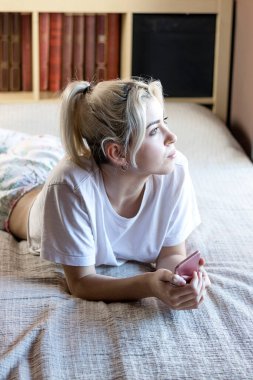 Rahat genç bir kadın cep telefonu tutarken yatakta uzanıyor.