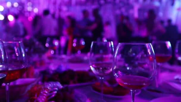 Una fiesta juvenil en un restaurante o un club nocturno, mesas de banquetes con alcohol y comida sobre el fondo de siluetas de personas bailando, escenario ligero y relleno púrpura, poca profundidad de campo — Vídeo de stock