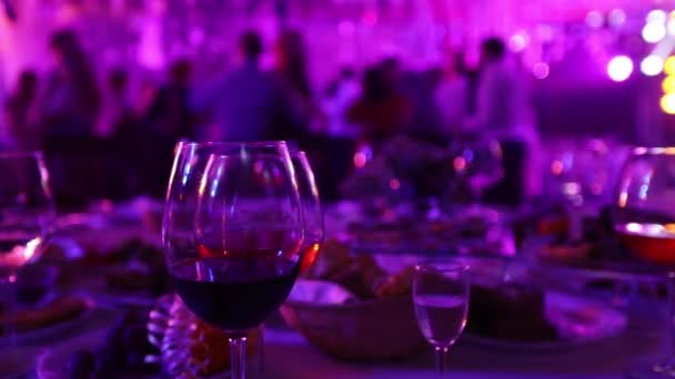Молодіжна вечірка в ресторані або нічному клубі, бенкетні столи з алкоголем і їжею на тлі силуетів танцюючих людей, сценічне світло-фіолетове наповнення, мала глибина різкості — стокове відео