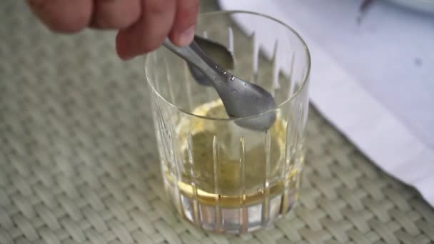 Chelnerul pune cuburi de gheață într-un pahar cu whisky, un pahar de whisky pe masă, un pahar de whisky pune cuburi de gheață, un pahar de whisky cu gheață pe masă — Videoclip de stoc