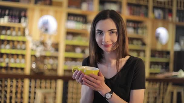 Girl in a restaurant with a cup of coffee, enjoying aroma and flavor of coffee while relaxing at coffee shop. Young beautiful happy smiling girl holding mug with hot coffee or tea — Stock Video