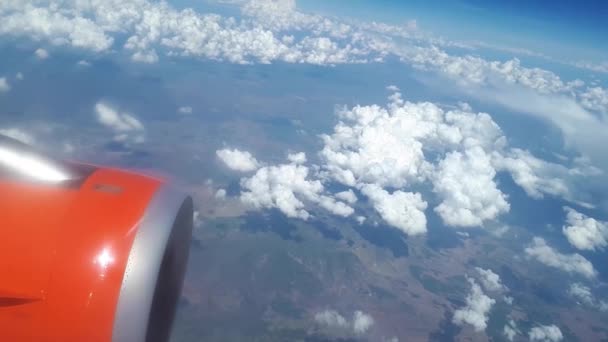 Vista desde la ventana del avión al cielo azul y nubes blancas, una turbina naranja en el ala del avión, una vista de la tierra desde el cielo a través de las nubes, nubes blancas flotan sobre el suelo — Vídeo de stock
