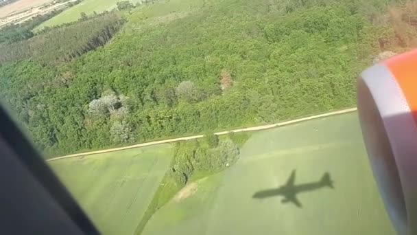 A sombra de um avião voador no chão num dia ensolarado. Vista da janela do avião. Sombra do avião. Voando sobre o campo e a floresta ao pousar no aeroporto — Vídeo de Stock