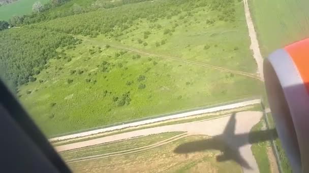 晴れた日に地面に飛行の機体の影。飛行機の窓からの眺め。平面の影。フィールドと空港に着陸中の森上空 — ストック動画