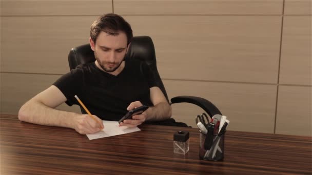Un joven trabaja en una mesa en la oficina. Un hombre de negocios escribe y dibuja en una hoja de papel blanco — Vídeos de Stock