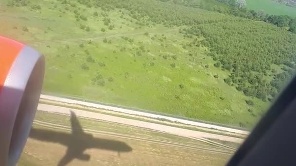 De schaduw van een vliegende vlak op de grond op een zonnige dag. Uitzicht vanuit vliegtuig raam. Schaduw van het vliegtuig. Vliegen over het veld en bos tijdens de landing op de luchthaven — Stockvideo
