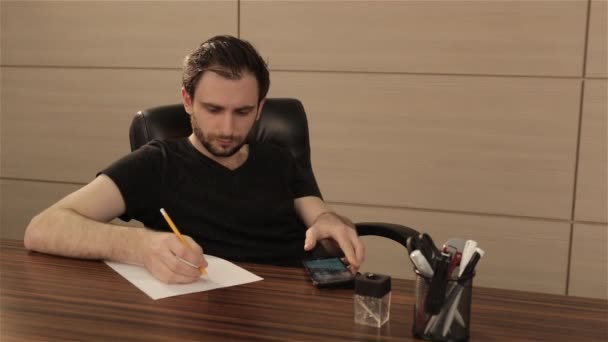 Un joven trabaja en una mesa en la oficina. Un hombre de negocios escribe y dibuja en una hoja de papel blanco — Vídeo de stock