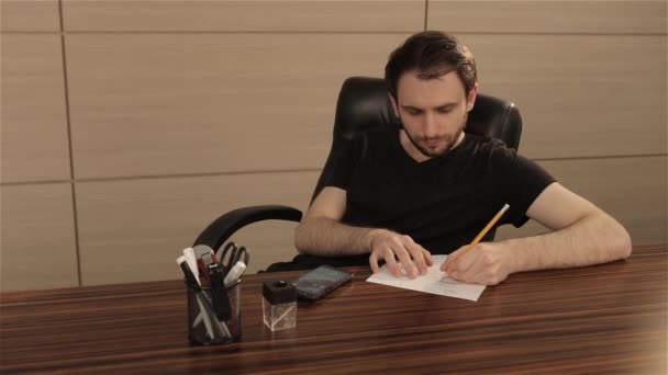 Um jovem está a trabalhar numa mesa no escritório. Um homem de negócios escreve e desenha em uma folha de papel branco — Vídeo de Stock