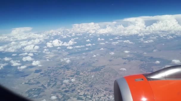 Uitzicht vanaf het vliegtuig venster naar de blauwe hemel en witte wolken, een oranje turbine op de vleugel van het vliegtuig, een weergave van de aarde vanuit de lucht door de wolken, witte wolken zweven boven de grond — Stockvideo
